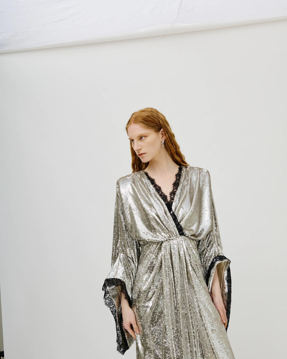 A model with red hair wearing a shimmering silver maxi dress with black lace trim and wide bell sleeves, standing against a plain white studio background.