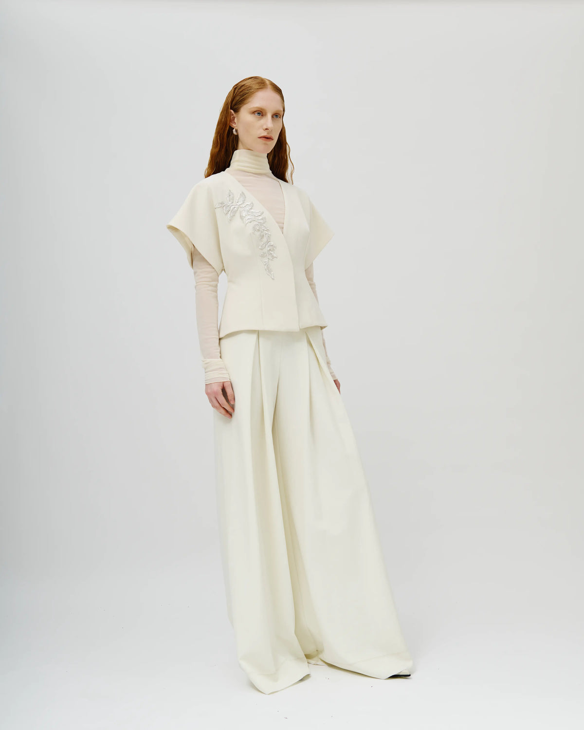 A model stands against a light gray studio background wearing a cream-colored Avaro Figlio two-piece outfit consisting of a short-sleeved cropped top with embroidery and matching wide-leg trousers.