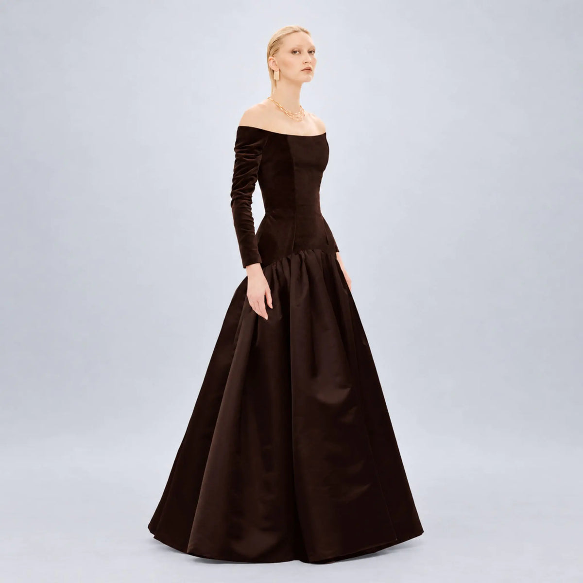 A woman modeling an off-the-shoulder Avaro Figlio long-sleeve ball gown in dark brown velvet and satin, photographed against a light gray studio background.