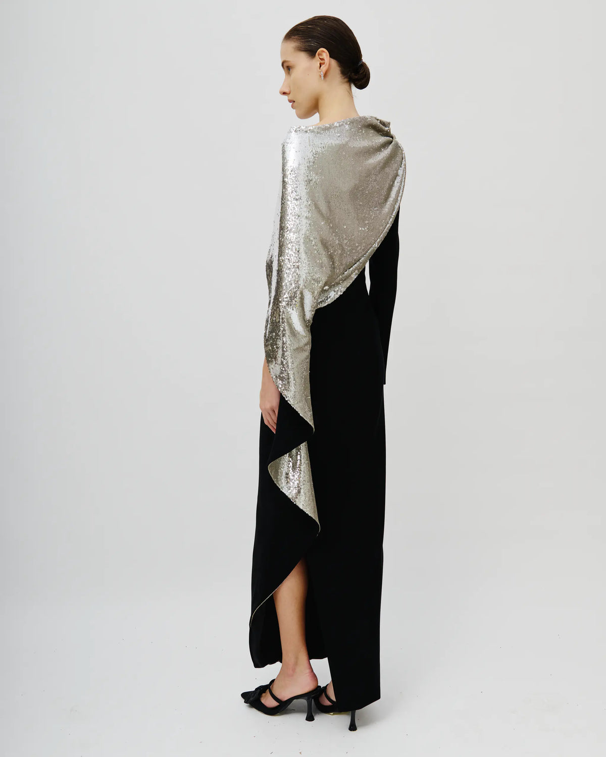 A model is shown from the back wearing a long black dress with a draped, asymmetrical silver sequined overlay, paired with black high-heeled sandals, against a plain light gray studio background.