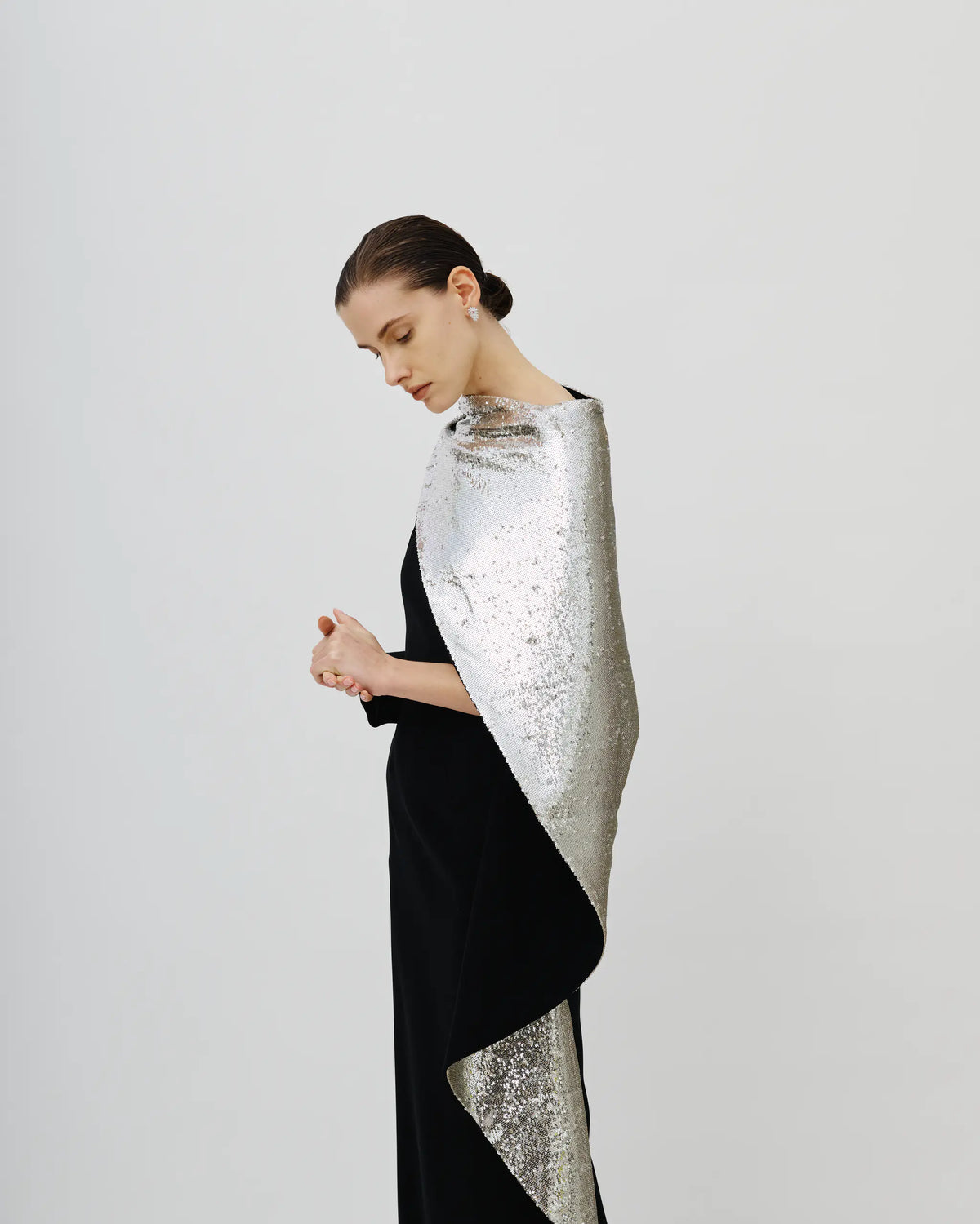 A model in a black dress wears a dramatic silver sequined wrap or shawl draped over her shoulders, posed against a solid light gray studio background.