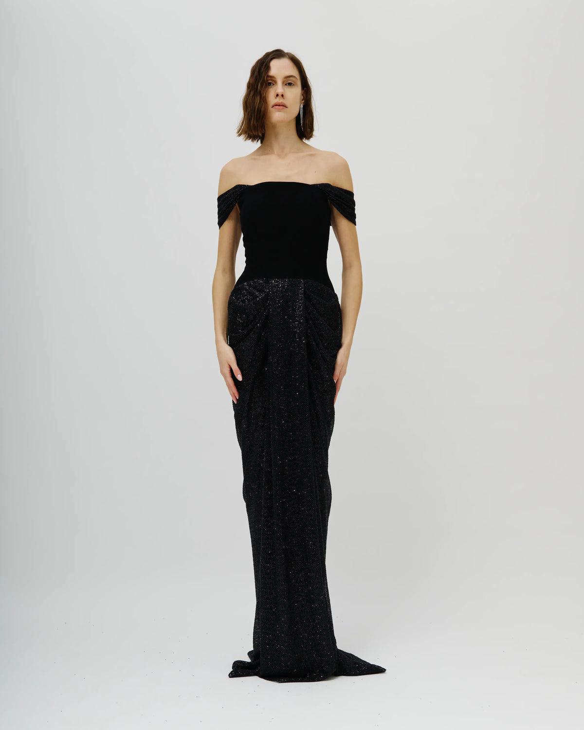 A model wearing a n Avaro Figlio floor-length black off-the-shoulder gown with a structured bodice and a heavily embellished, textured skirt, standing against a light gray studio background.