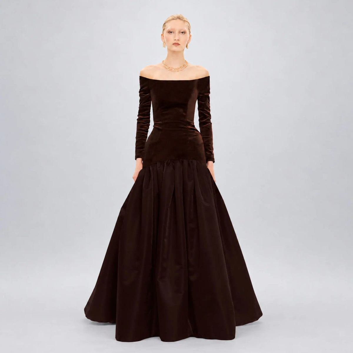 Model wearing an off-the-shoulder Avaro Figlio long-sleeve ball gown in dark brown velvet and satin, photographed against a light gray studio background.