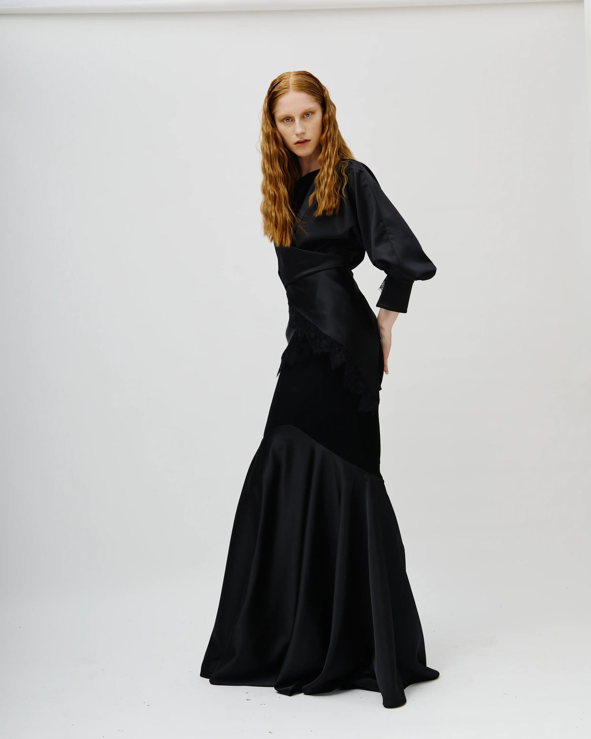 Model with long red hair wearing a black Avaro Figlio satin mermaid gown with dramatic puffed sleeves, standing against a plain white studio background.