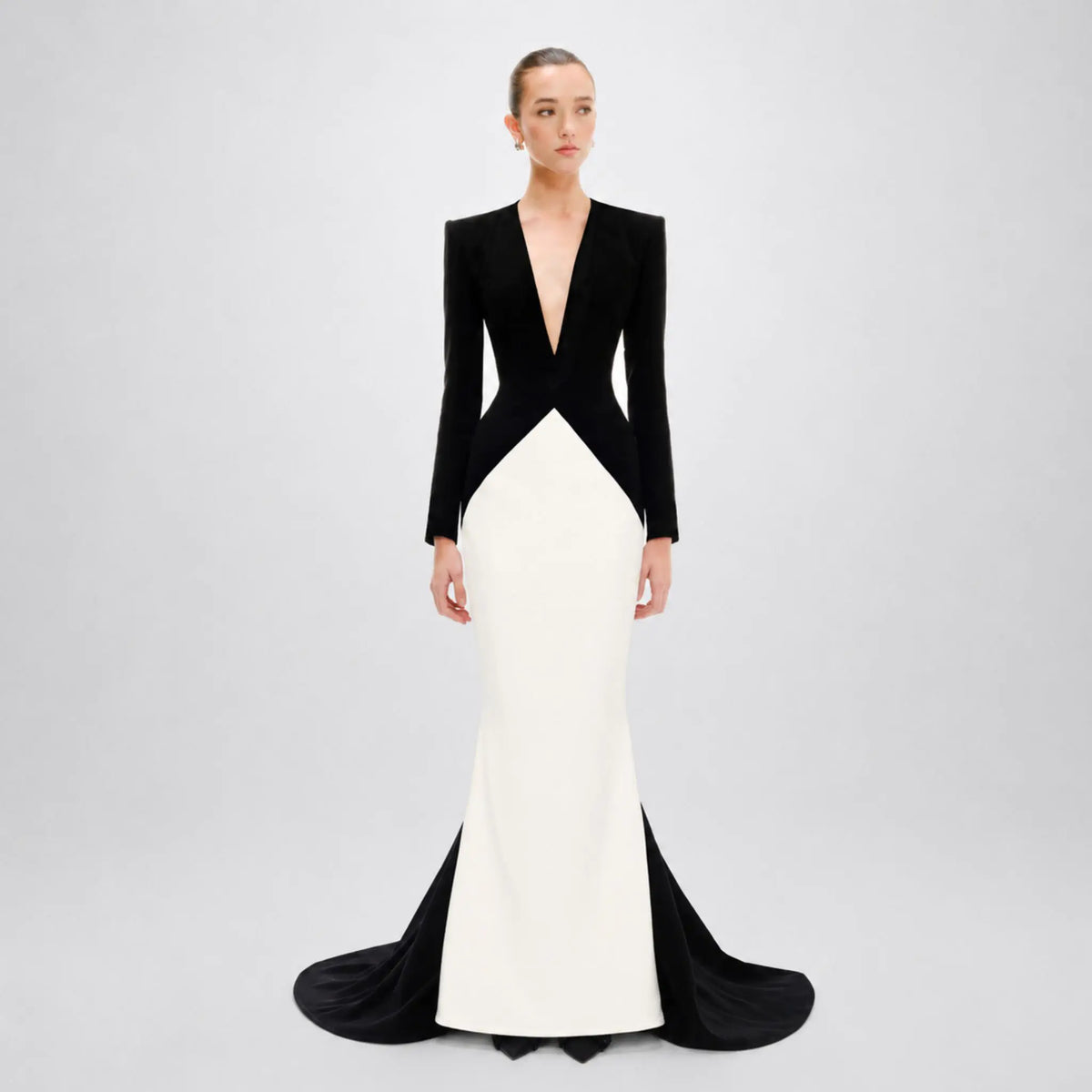 A model wearing a long-sleeved black and white Avaro Figlio mermaid gown with a deep V-neckline and structured shoulders, set against a plain light gray studio background.