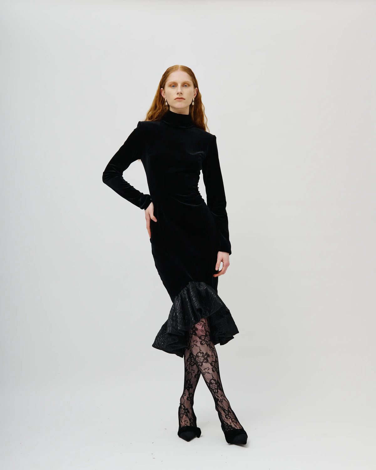 A model with red hair wearing a black Avaro Figlio velvet long-sleeve dress with a ruffled hem, paired with black lace tights and black heels, photographed against a solid light gray background.