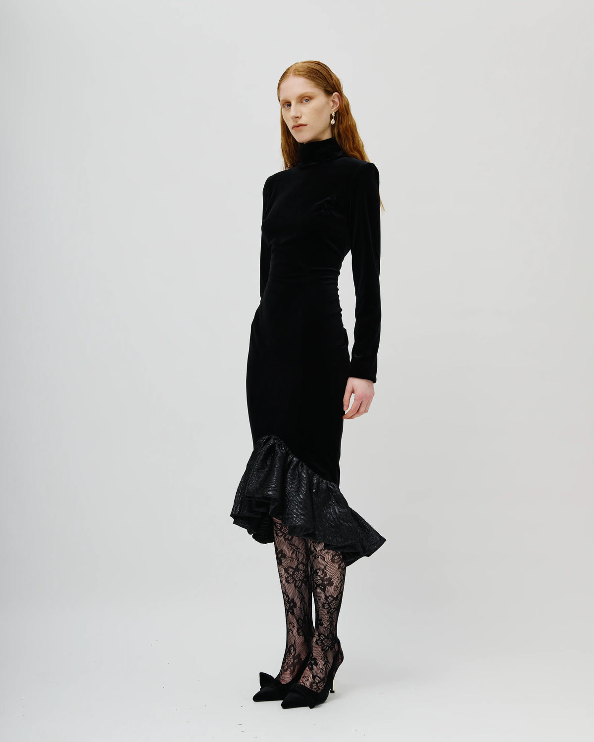 A model with red hair is wearing a black Avaro Figlio velvet long-sleeve turtleneck dress with a ruffled hem, paired with black lace tights and black pointed-toe heels, photographed against a light gray studio background.