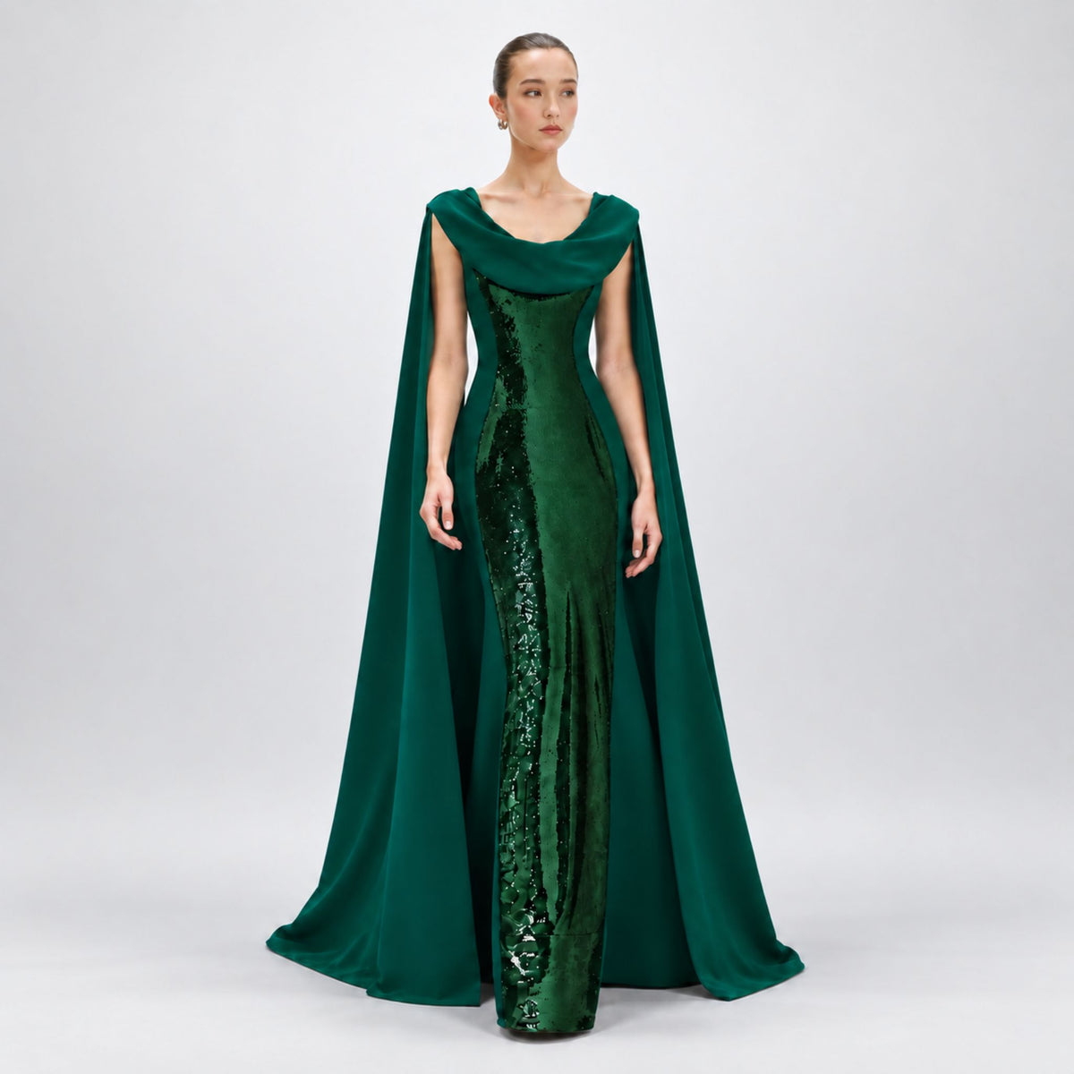 A model wearing a floor-length Avaro Figlio forest green gown with a draped cape and a vertical sequined panel down the front, photographed against a light gray studio background.