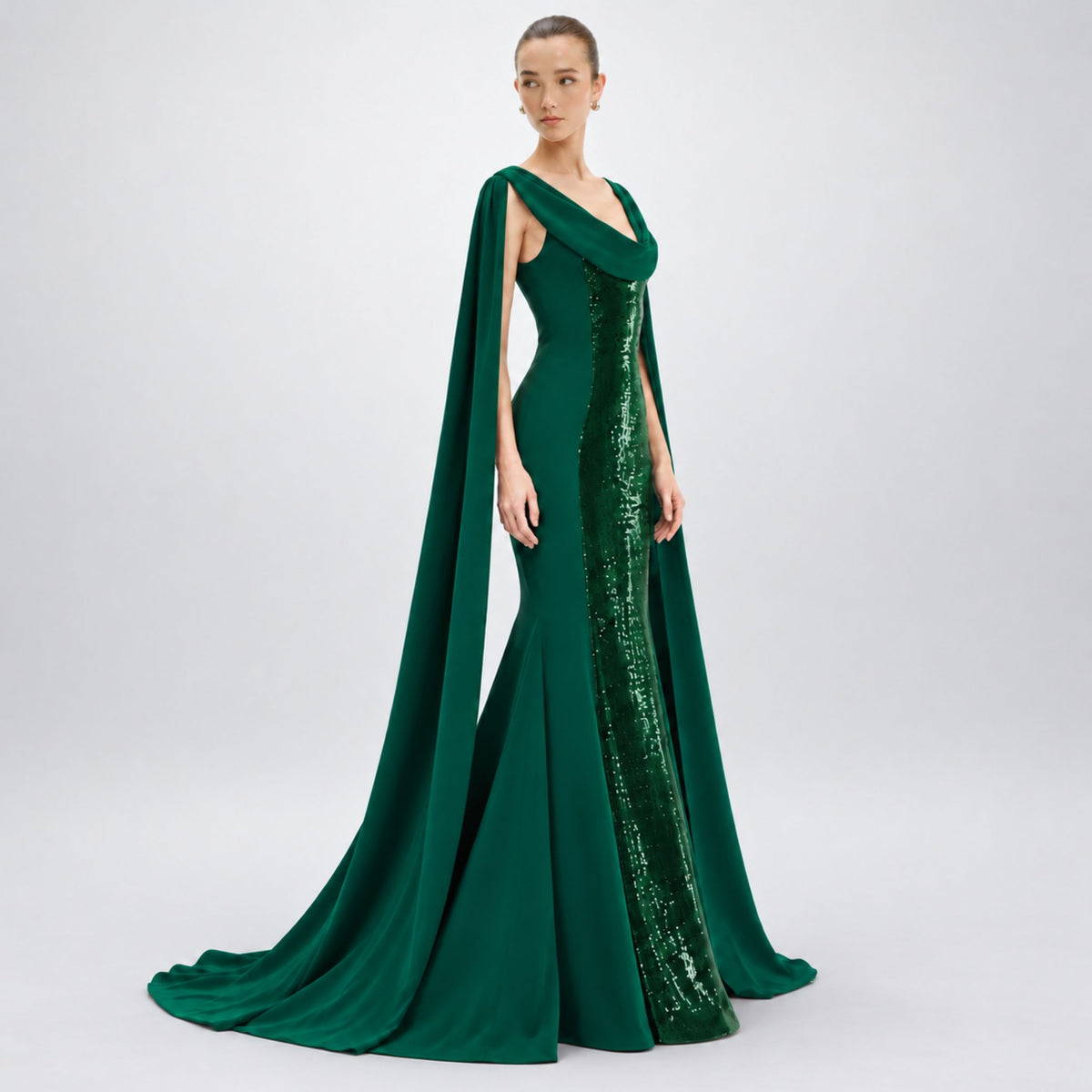 A model wears a floor-length Avaro Figlio forest green gown with a draped cape and a shimmering sequined side panel, photographed against a light gray studio background.