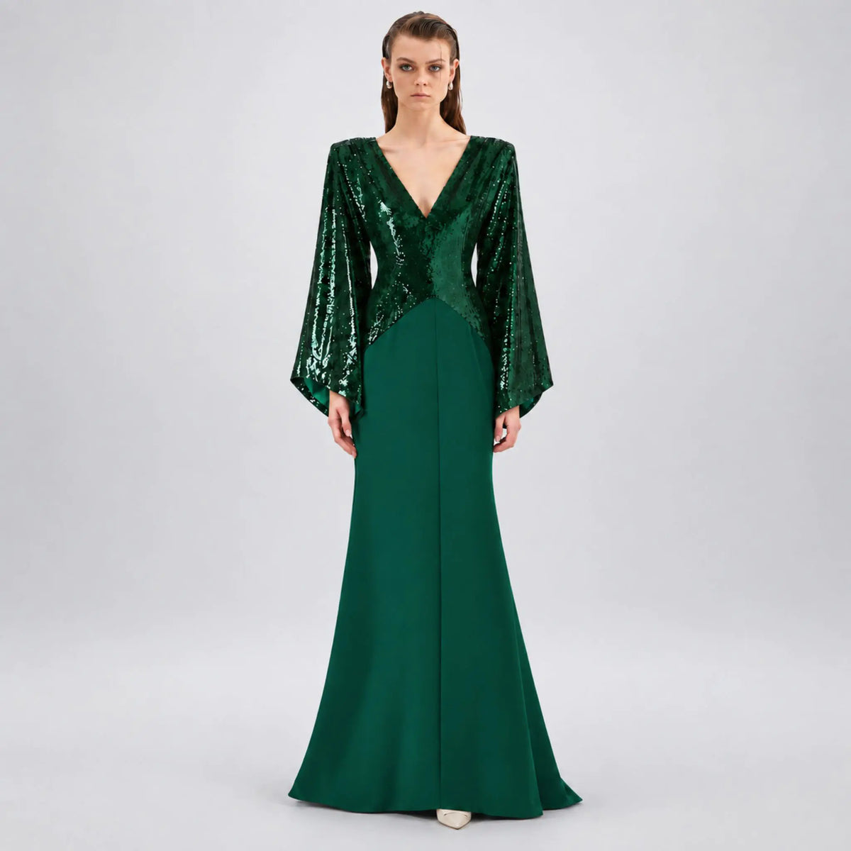 A model wearing a floor-length avaro figlio forest-green kimono maxi dress with a sequined bodice, deep V-neckline, and dramatic bell sleeves, photographed against a neutral light gray studio background.