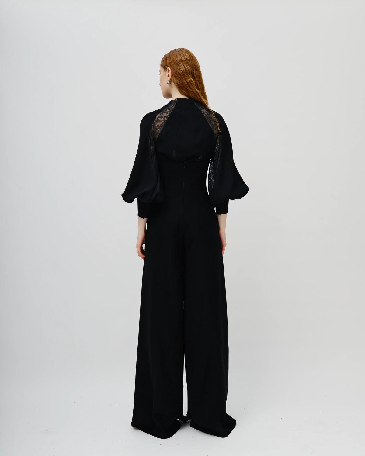 A model viewed from the back wearing a black Avaro Figlio wide-leg jumpsuit with sheer black lace shoulder panels, set against a plain light gray studio background.