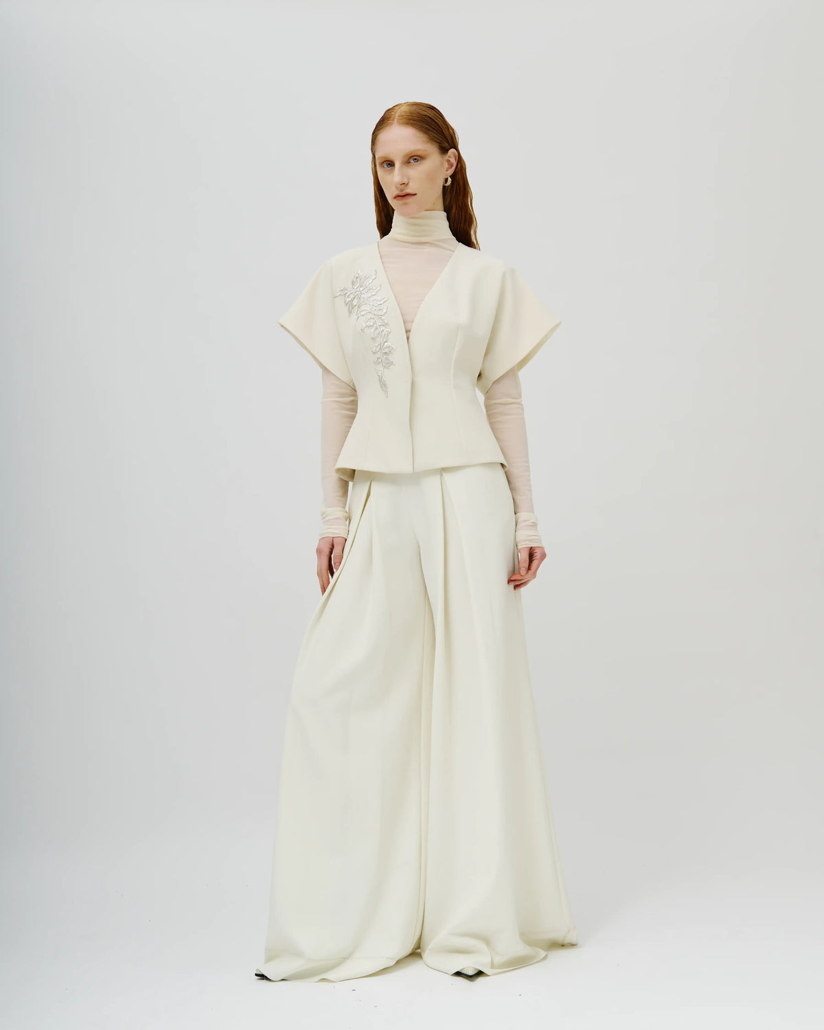 Model stands against a light gray studio background wearing a cream-colored Avaro Figlio two-piece outfit consisting of a short-sleeved cropped jacket with floral embroidery on the chest and matching wide-leg trousers.