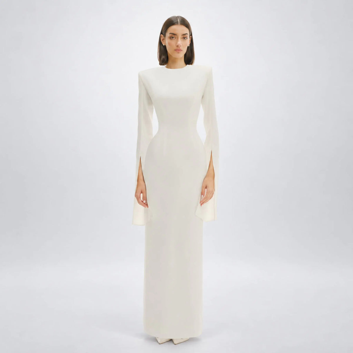 A model wearing a floor-length, white long-sleeve avaro figlio gown with flared bell sleeves and side slit details, standing against a seamless light gray studio background.