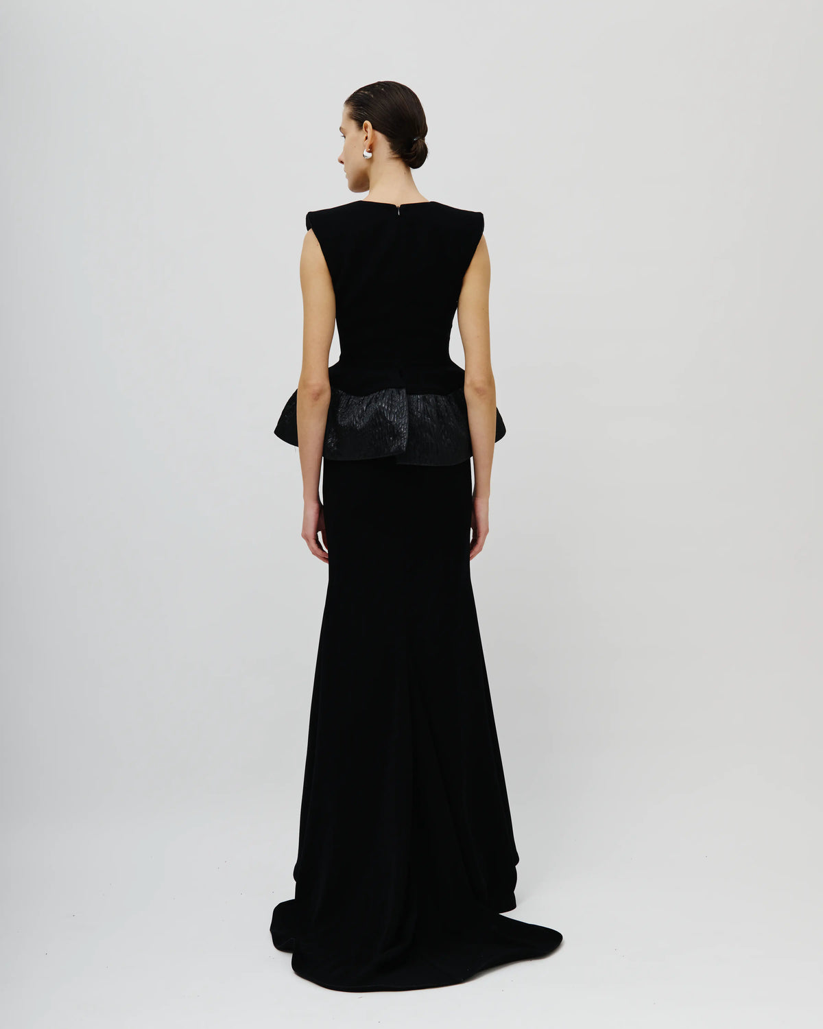 A model viewed from the back wearing an Avaro Figlio floor-length black evening gown with a structured velvet-like bodice, peplum-like waist detail, and flared skirt on a light gray studio background.