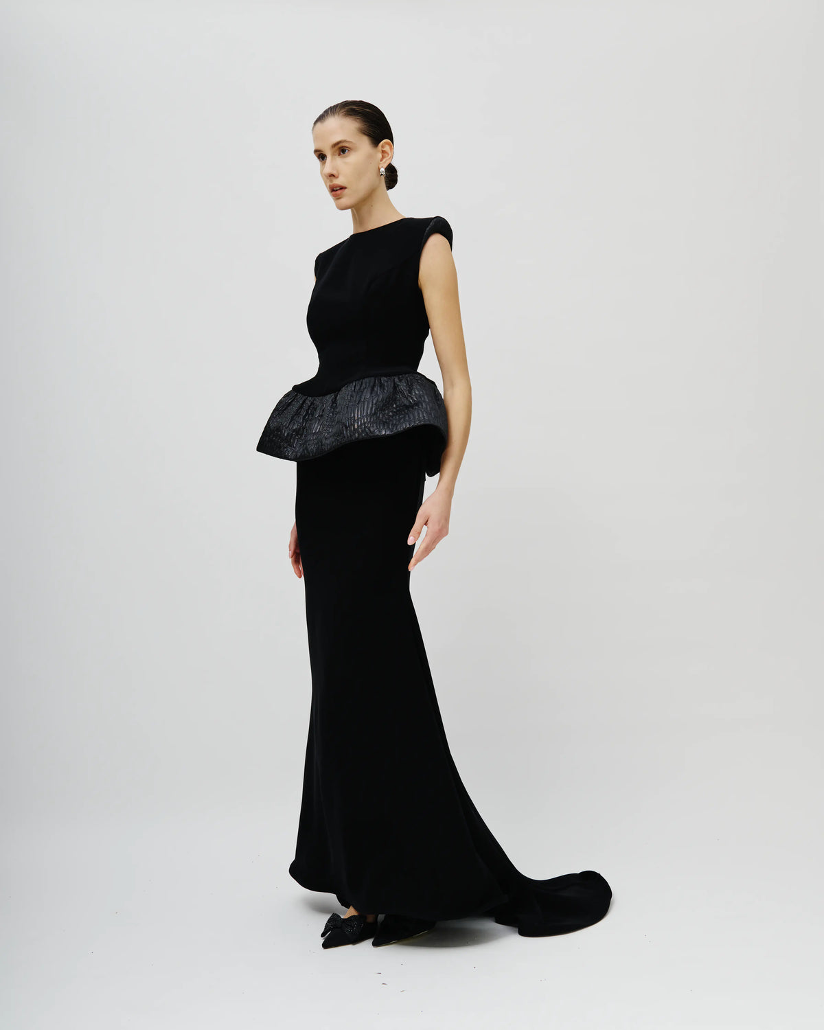A model wearing an Avaro Figlio black evening gown with a textured peplum waist, photographed against a seamless light gray studio background.