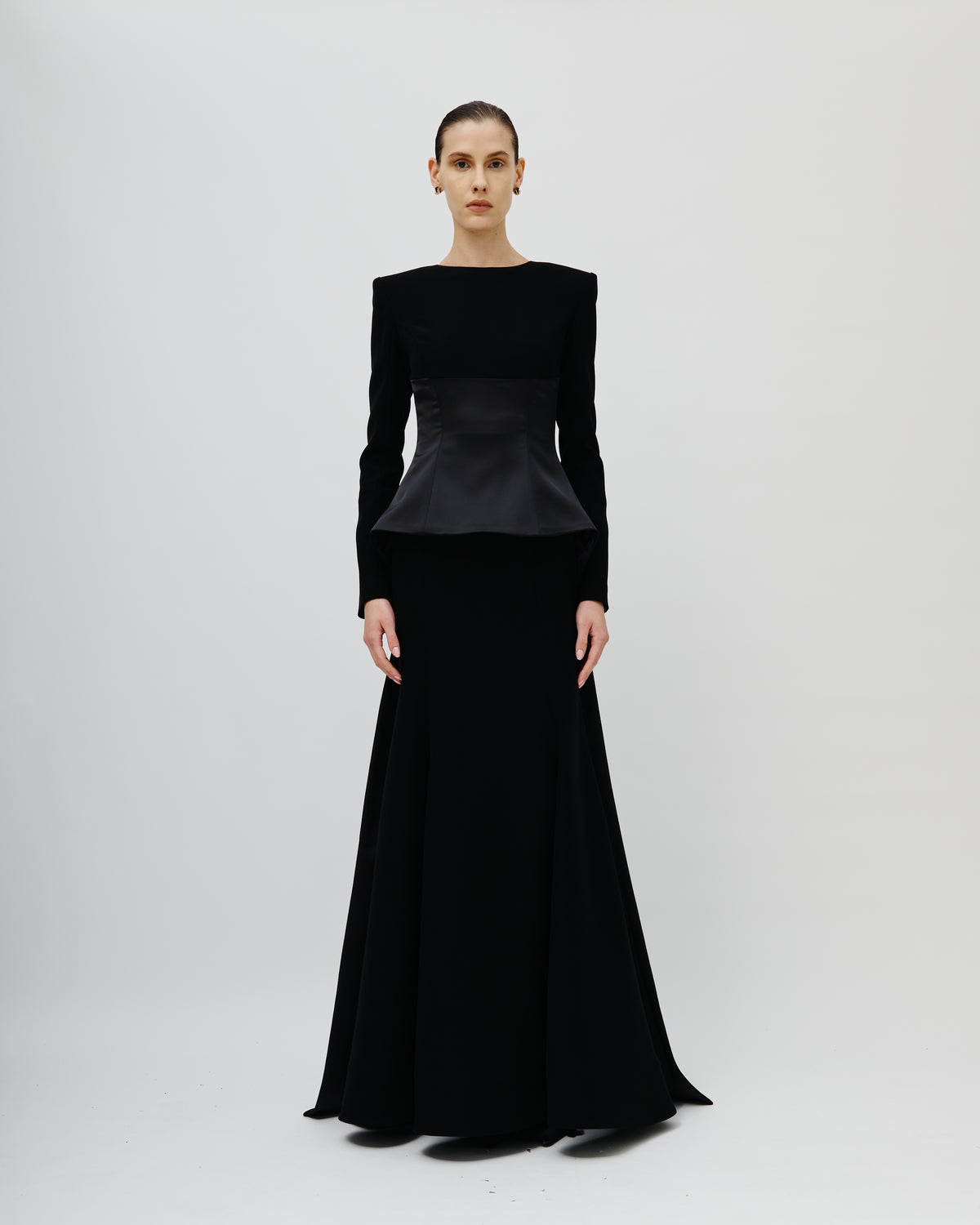 A model stands against a light gray studio background wearing an Avaro Figlio long black evening gown with long sleeves, structured shoulders, a peplum waist, and a full floor-length skirt.