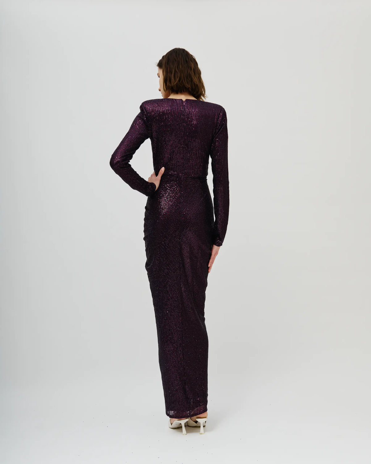 A woman viewed from the back wearing an Avaro Figlio long-sleeved, floor-length deep purple sequined gown against a plain light gray studio background.