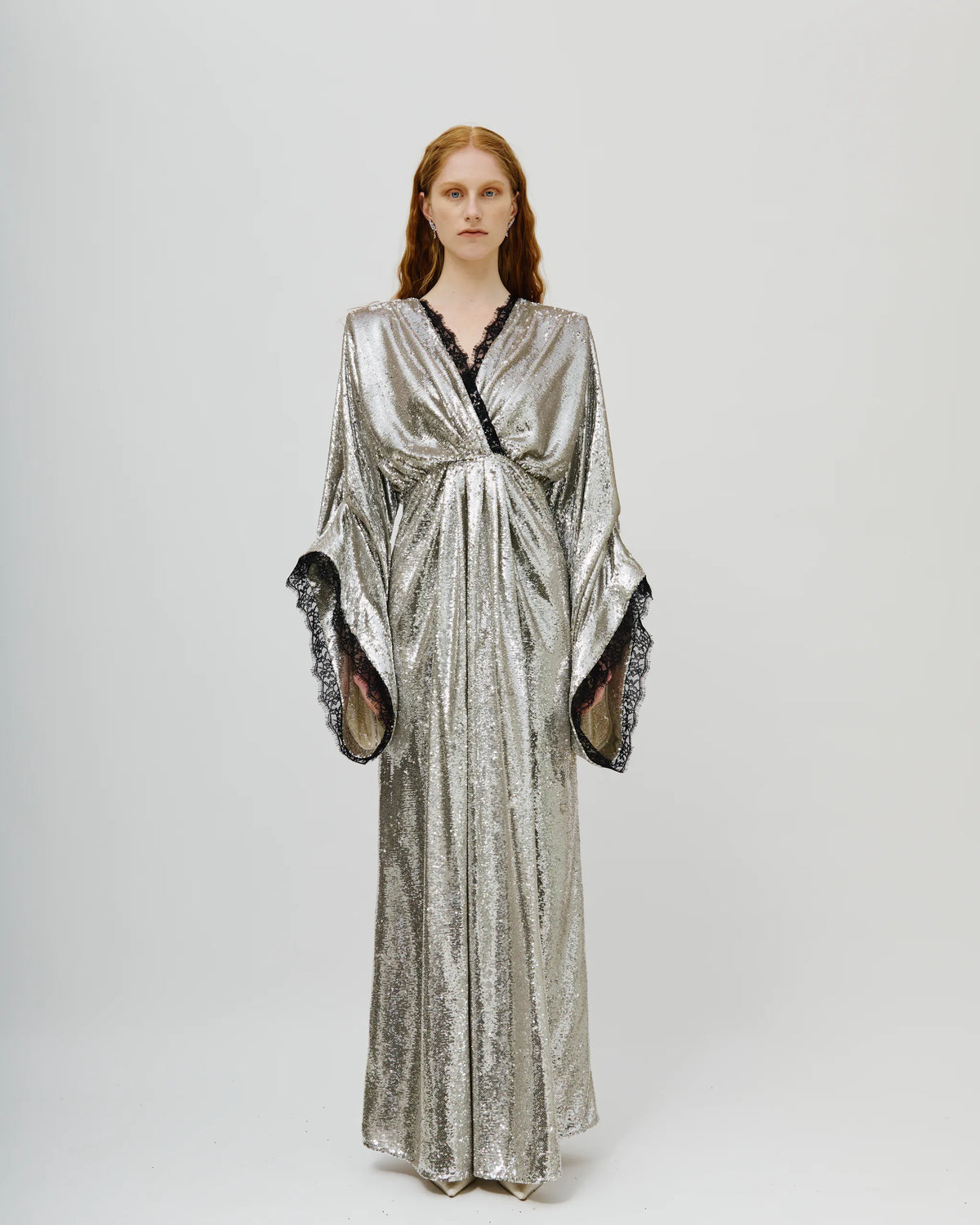 A model wearing a long, shimmering metallic silver wrap dress with wide draped sleeves and black lace trim, standing against a light gray studio background.