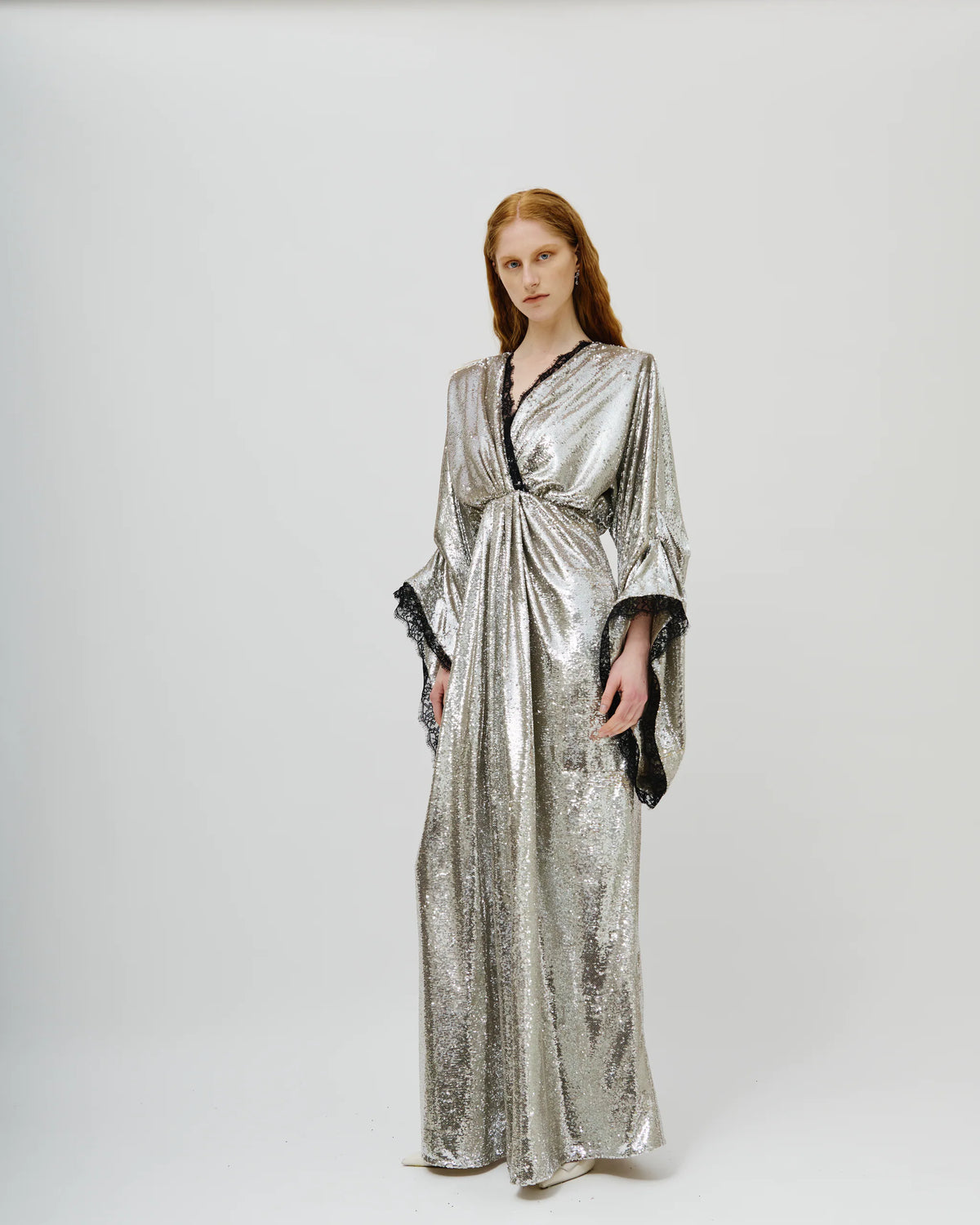 A model wearing a floor-length, shimmering silver robe with black lace trim and wide draped sleeves, standing against a plain light gray studio background.