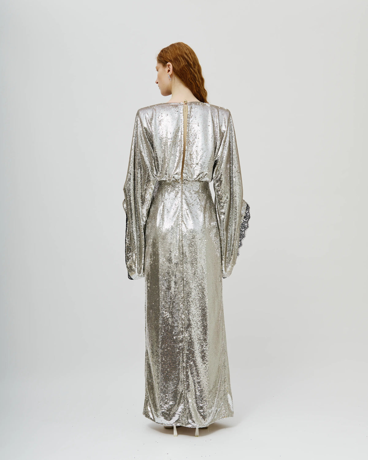 A woman with red hair is shown from the back wearing a floor-length, long-sleeved metallic silver gown with a textured, sequined surface and a back zipper, set against a plain light gray studio background.
