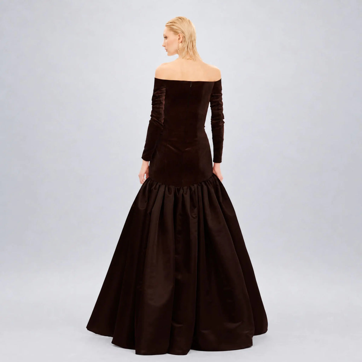 A woman modeling an off-the-shoulder dark brown  avarofiglio velvet ball gown with long sleeves and a full skirt, shown from the back against a light gray studio background.