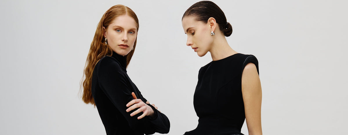 Two models wearing elegant black evening gowns in a studio setting. The model on the left wears a long-sleeved black velvet dress with a textured ruffled hem and black lace tights.