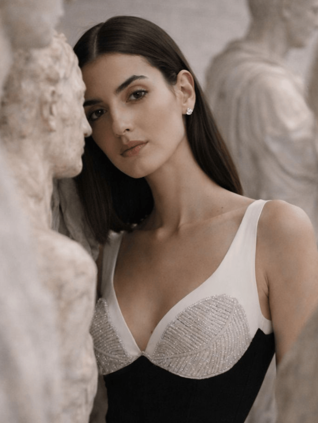 A woman wearing a black and white bustier-style top with textured beaded cups, posed between classical stone statues in a softly lit indoor setting.