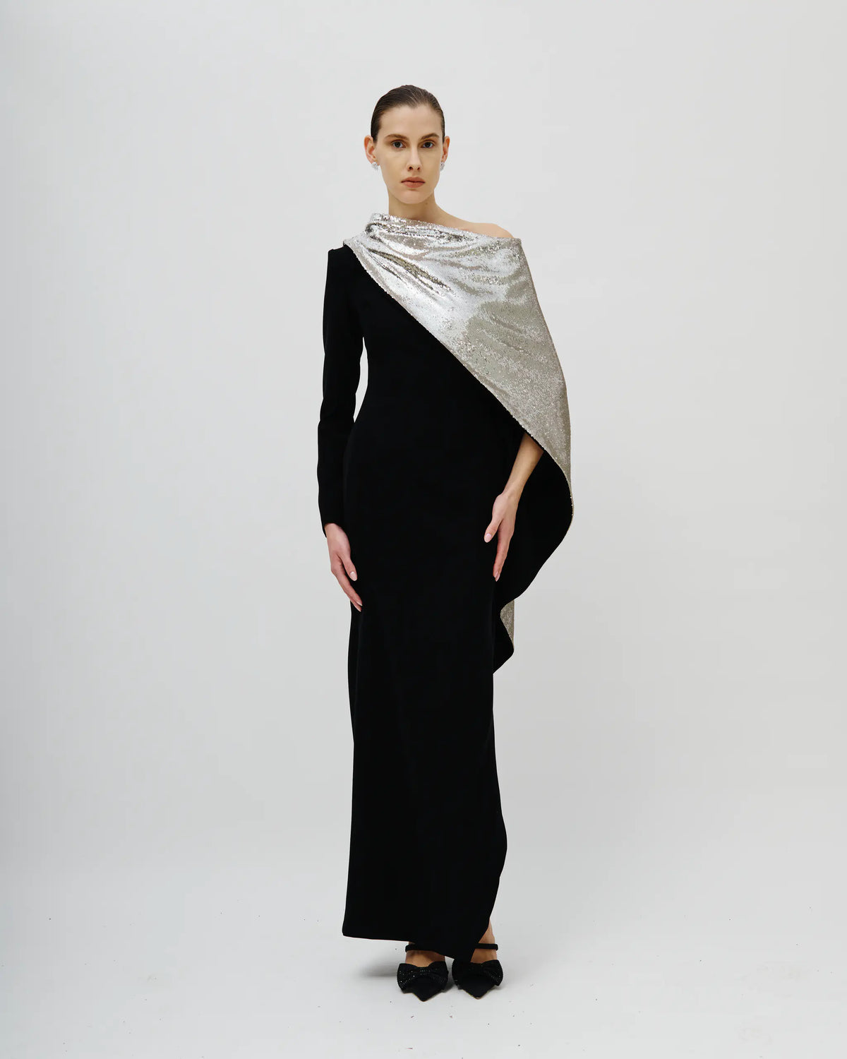 A model stands against a light gray studio background wearing a long black off-the-shoulder dress with a metallic silver draped shawl over one shoulder and black pointed shoes.