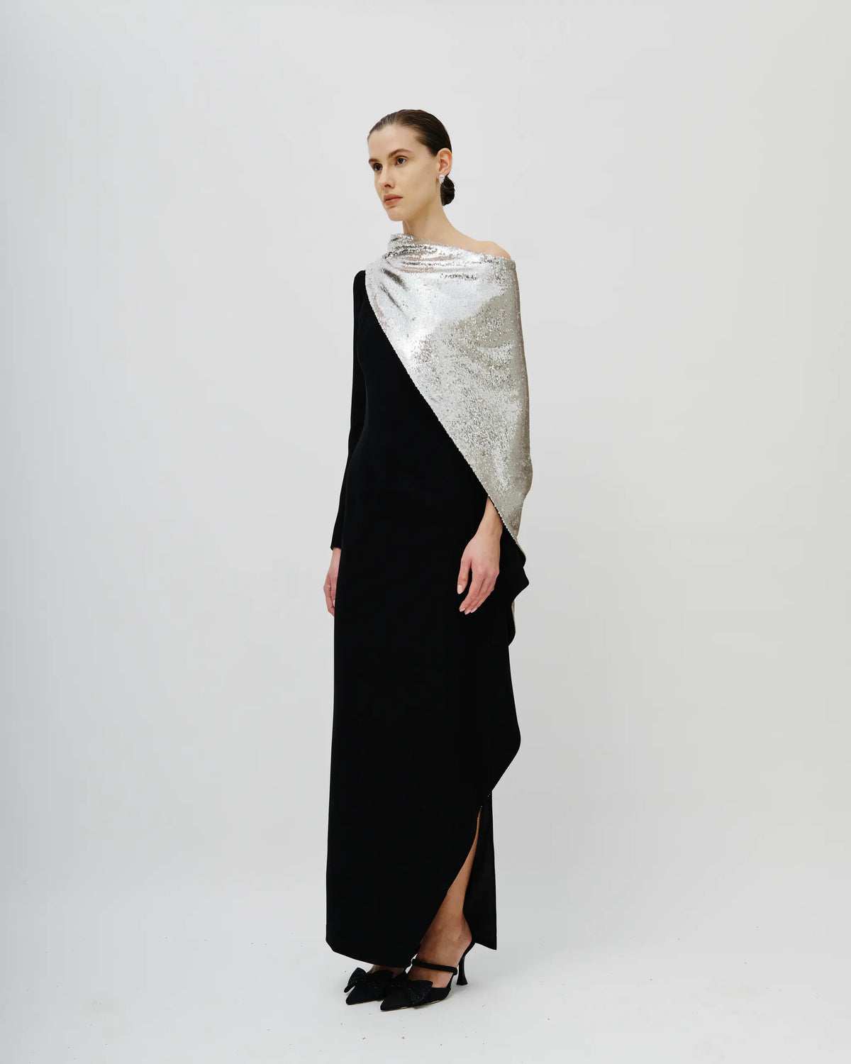 A model stands in profile wearing a long black dress with a draped silver sequined shawl over one shoulder, photographed against a plain light gray studio background.