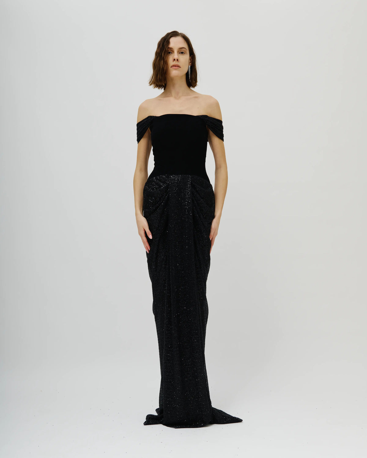 A model wearing a n Avaro Figlio floor-length black off-the-shoulder gown with a structured bodice and a heavily embellished, textured skirt, standing against a light gray studio background.