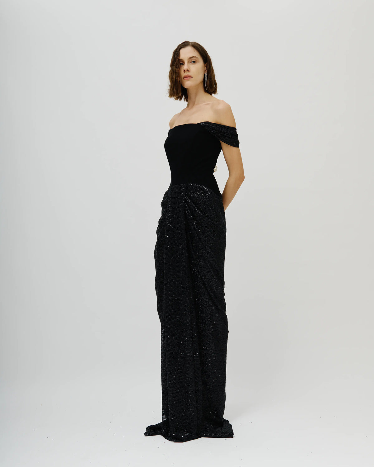 A woman modeling an Avaro Figlio off-the-shoulder black evening gown with a structured bodice and draped, textured skirt, photographed against a plain light gray studio background.