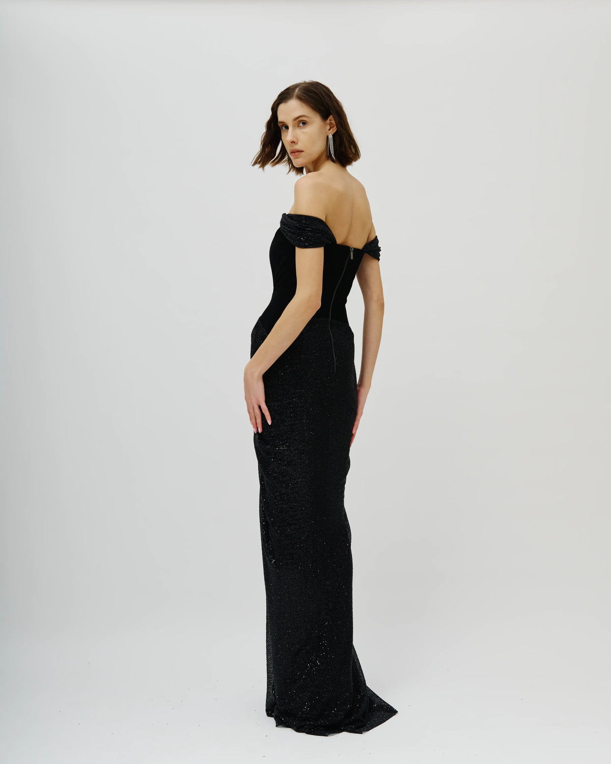 A woman modeling an Avaro Figlio floor-length off-the-shoulder black evening gown with a fitted silhouette and subtle shimmer, photographed against a plain light gray studio background.