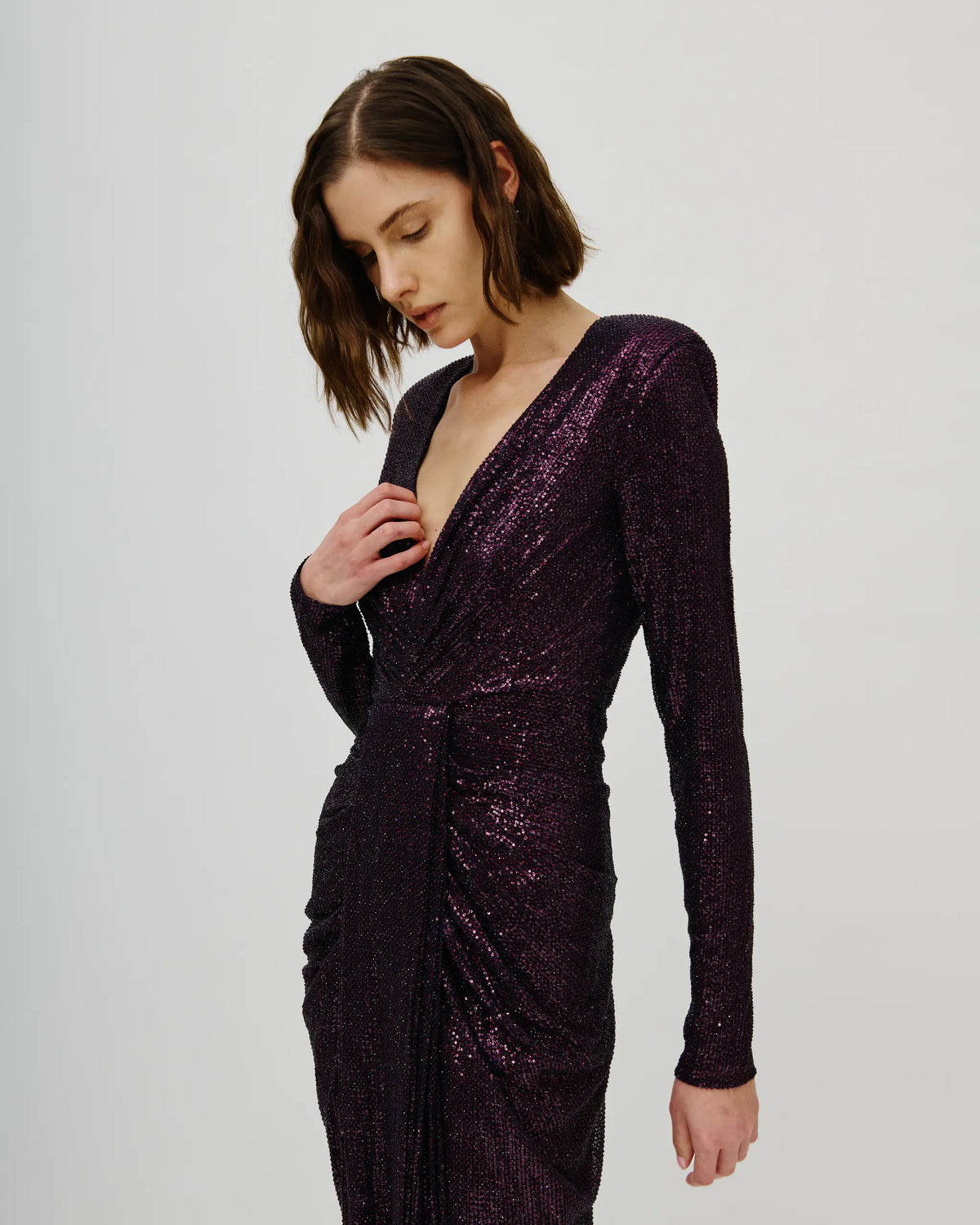 A woman wearing an Avaro Figlio long-sleeve, deep V-neck, ruched sequin dress in a dark plum purple, photographed against a plain light gray studio background.