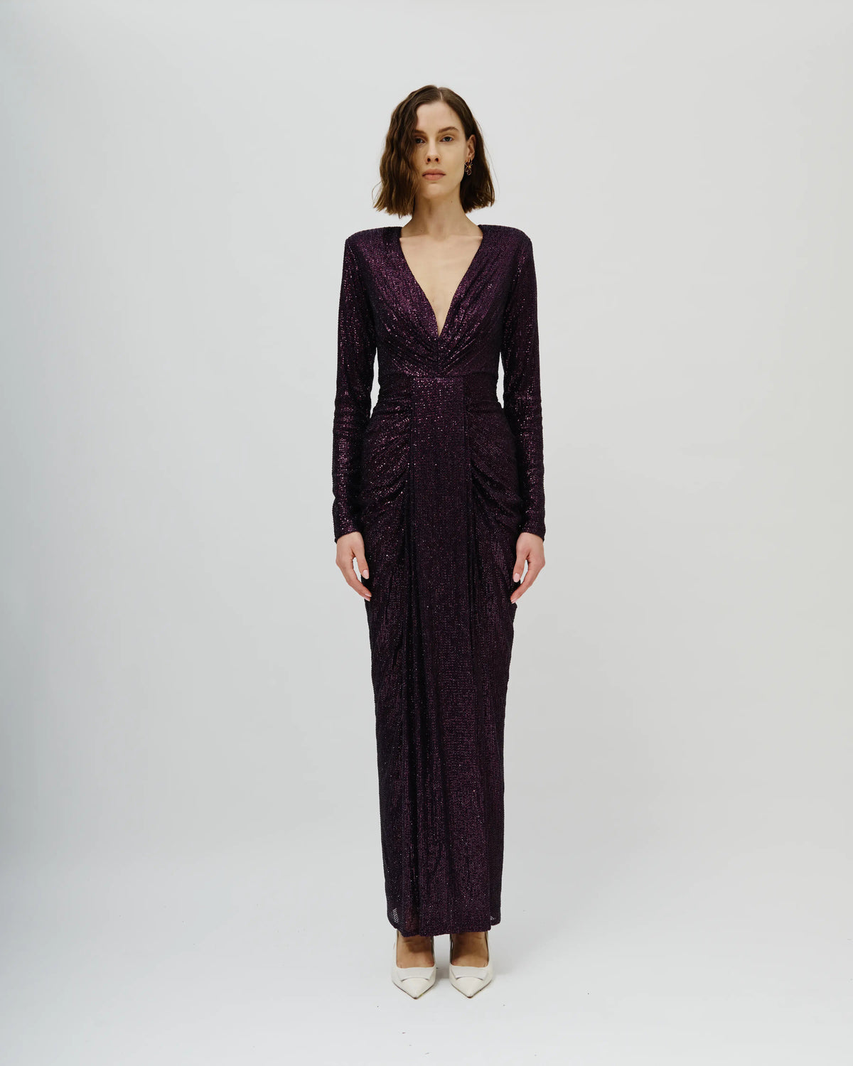 A woman modeling an Avaro Figlio long-sleeved, deep V-neck maxi dress in a textured, deep plum purple fabric, standing against a light gray studio background.