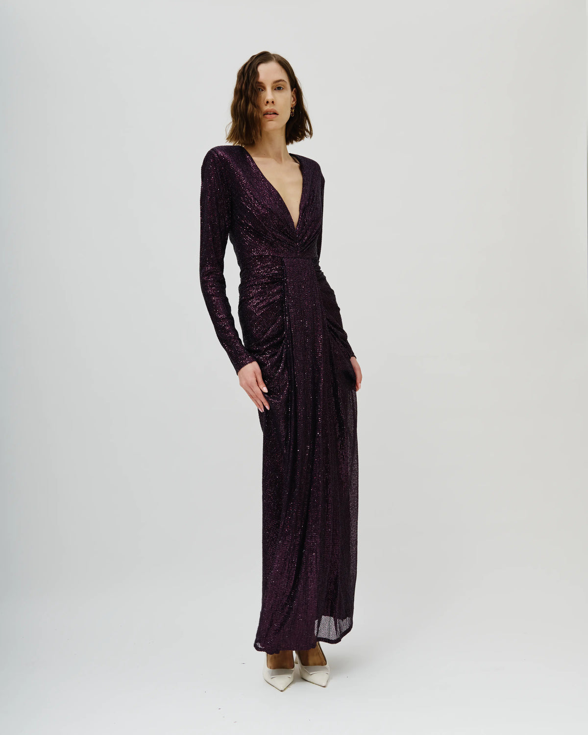 A woman modeling an Avaro Figlio long-sleeved, deep V-neck maxi dress covered in sparkling dark plum sequins, standing against a plain light gray studio background.