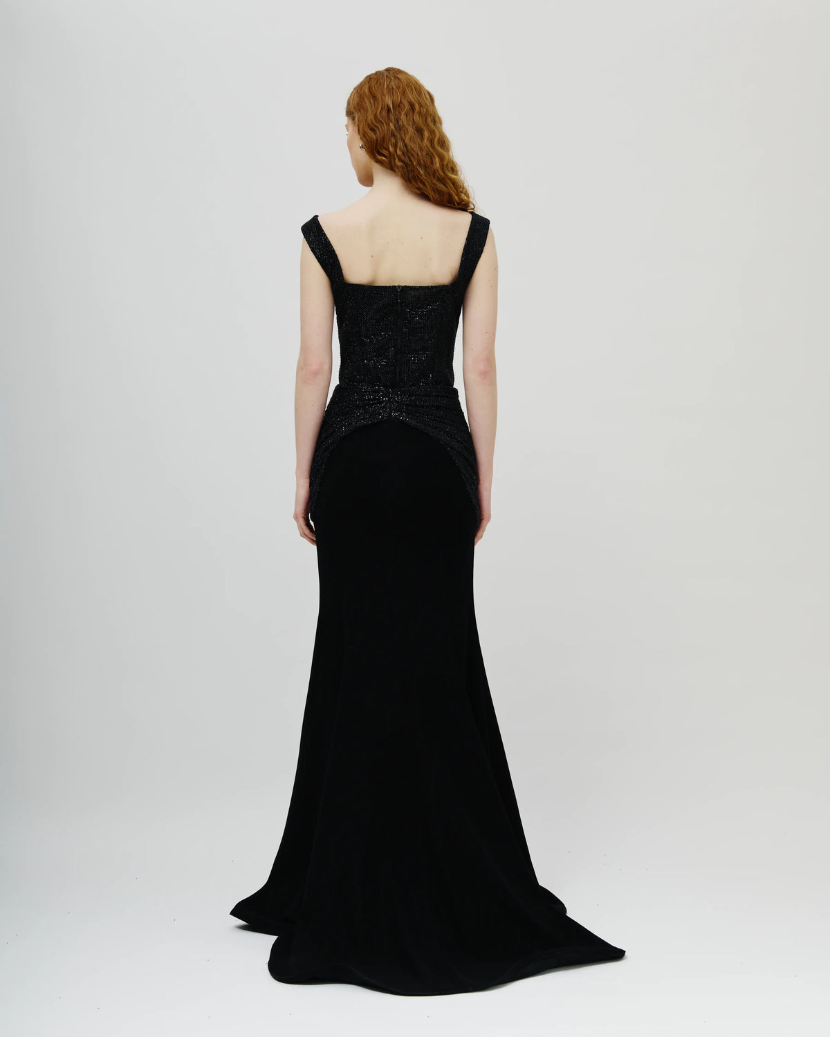 A woman with red hair is shown from the back Avaro Figlio wearing a floor-length black gown with a structured lace bodice and wide shoulder straps, photographed against a plain light gray studio background.