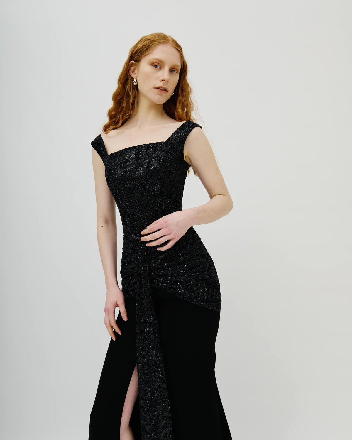 A woman with red hair wearing a black Avaro Figlio one-shoulder evening gown with a textured bodice and draped skirt, standing against a light gray studio background.