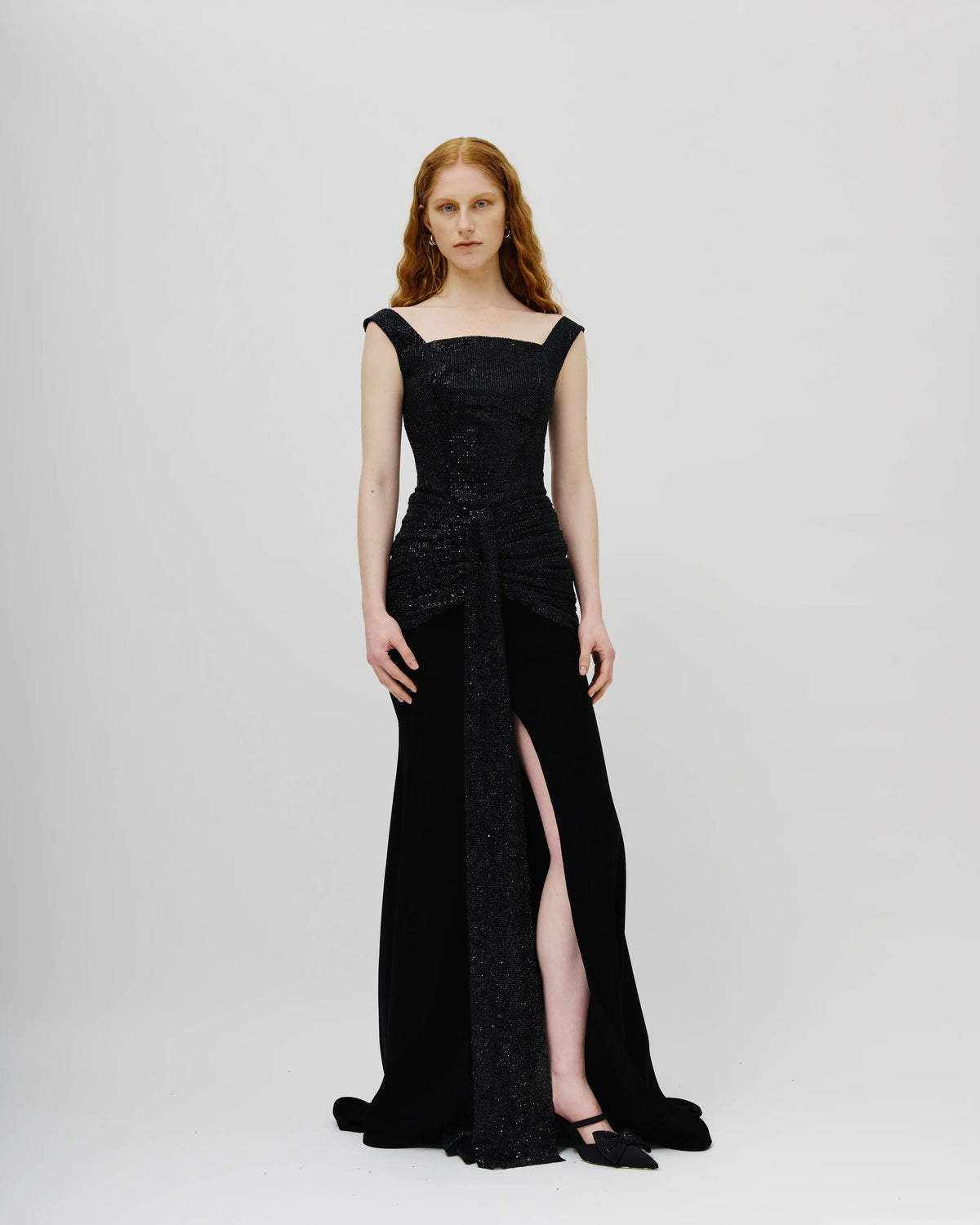 A model with red hair wearing a floor-length black Avaro Figlio evening gown with a high thigh slit, structured bodice, and draped train, standing against a light gray studio background.