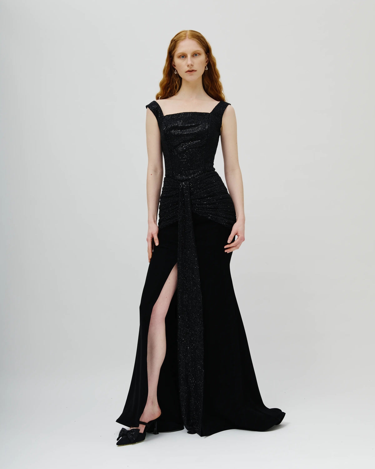 A model with red hair wearing a floor-length black Avaro Figlio evening gown with a structured bodice, draped waist detail, and a high thigh slit, photographed against a light gray studio background.