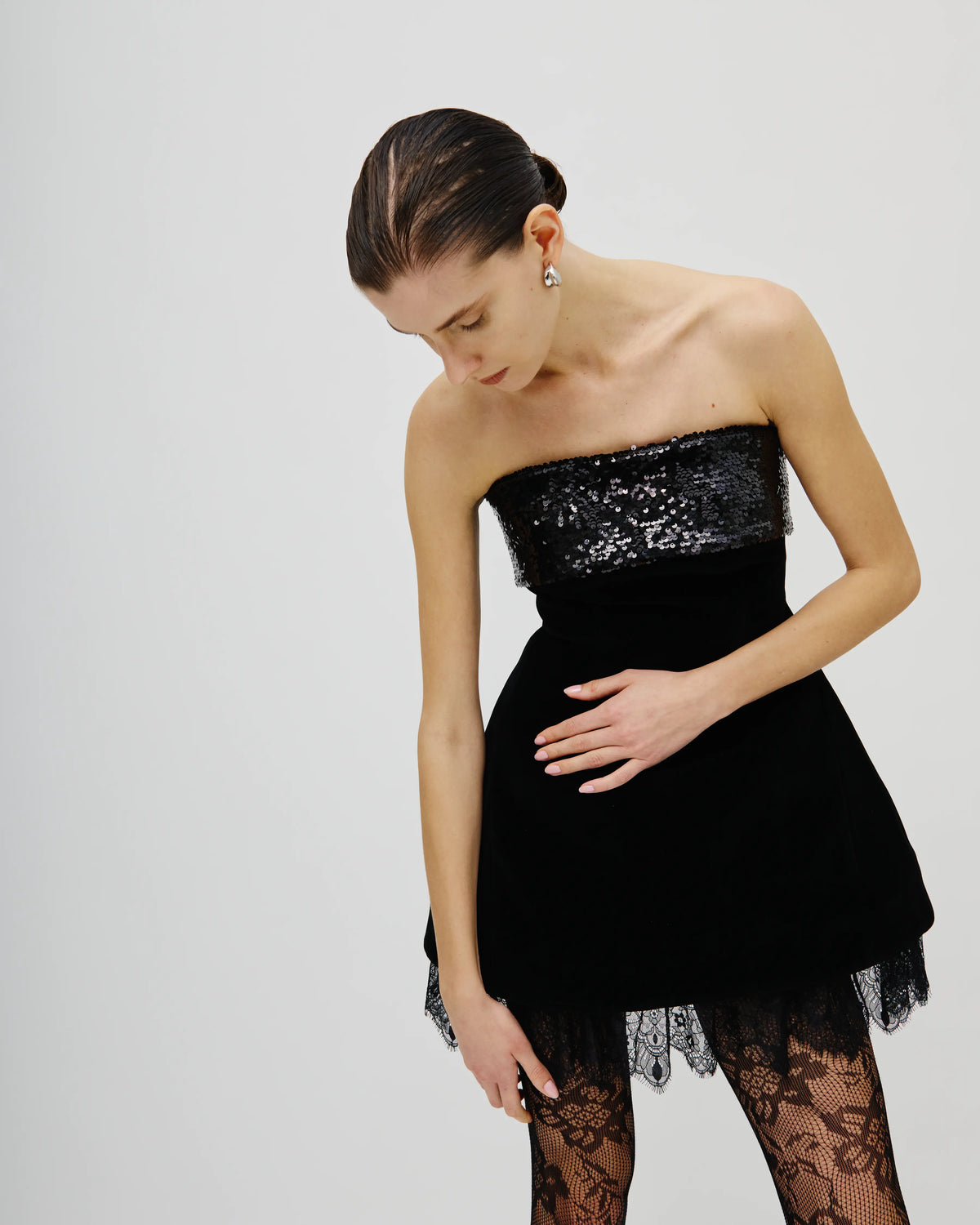 A model wearing a strapless black Avaro Figlio velvet mini dress with a sequined bodice, black lace tights, and a small silver earring, photographed against a light gray studio background.