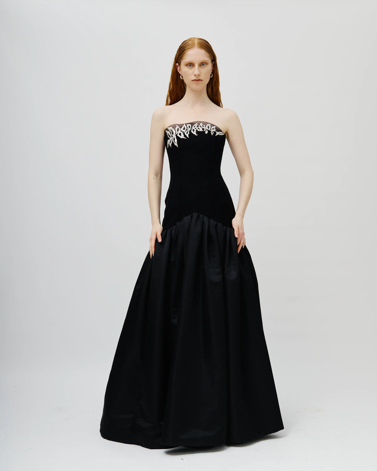 A model with red hair wearing an Avaro Figlio black strapless evening gown with silver embellishments on the bodice, standing against a light gray studio background.