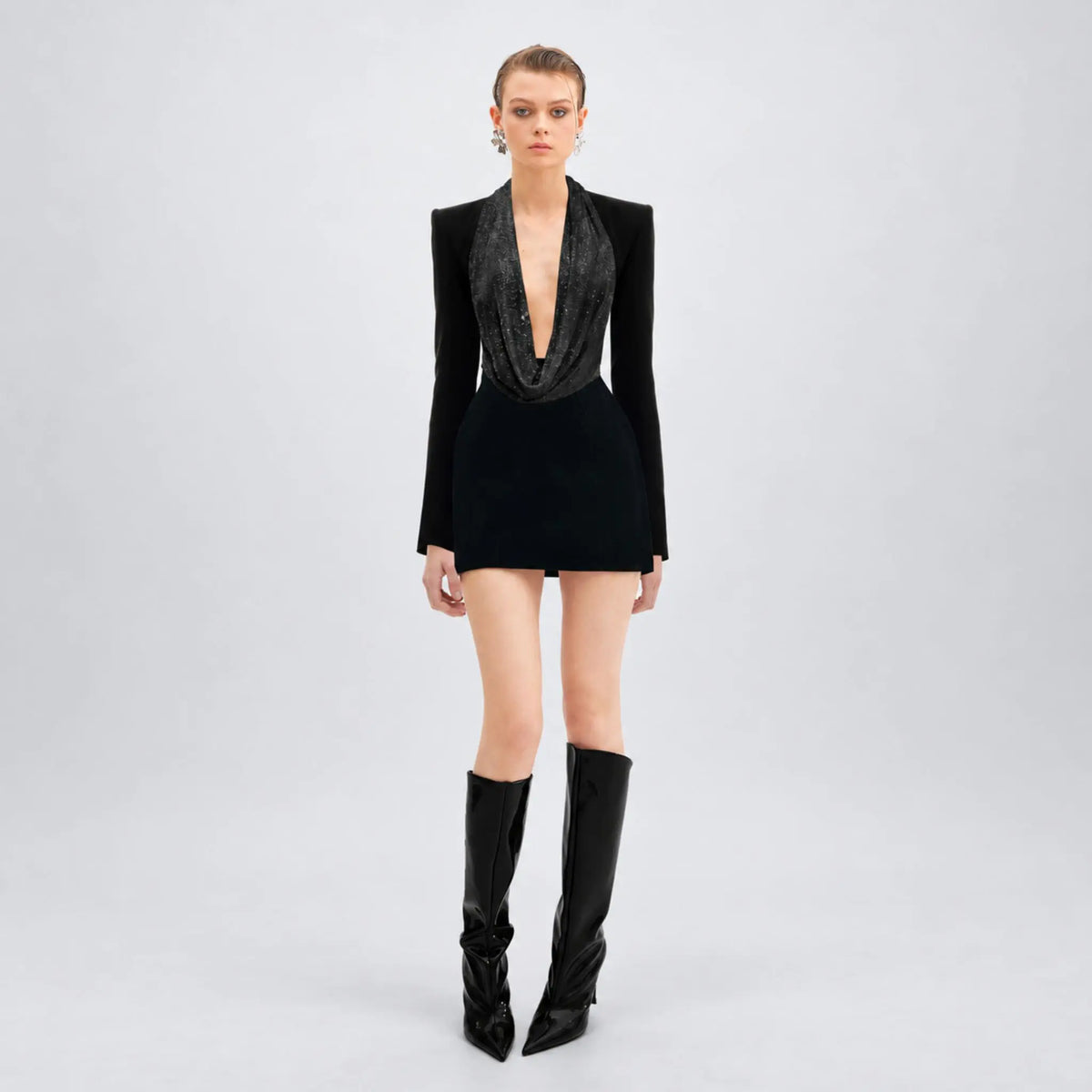 A model stands against a light gray studio background wearing a black avaro Figlio long-sleeve mini dress with a deep plunging cowl neckline, paired with black knee-high pointed-toe boots.