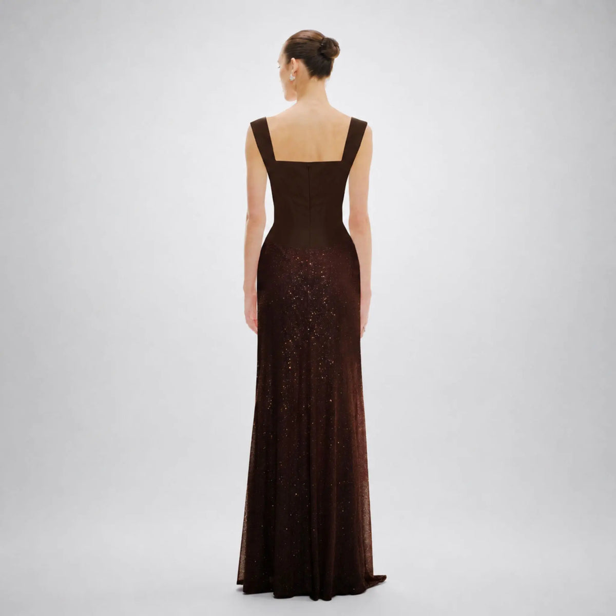 A woman stands with her back to the camera wearing a floor-length Avaro Figlio dark brown evening gown with a square neckline and subtle shimmering sequin detailing on the skirt, photographed against a light gray studio background.