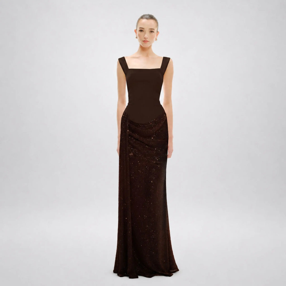 A model wearing a floor-length, dark brown Avaro Figlio evening gown with a square neckline and shimmering sequined detailing on the skirt, set against a plain light gray studio background.