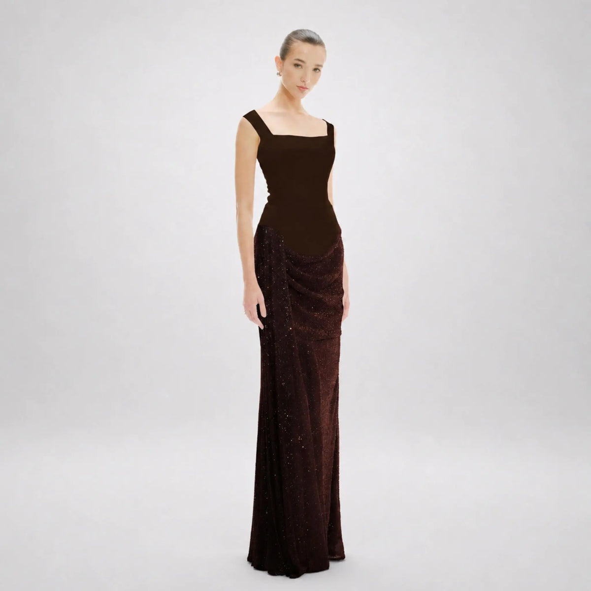 A model wearing a floor-length avarofiglio dark brown evening gown with a structured square neckline, fitted bodice, and draped sequined skirt, photographed against a light gray studio background.