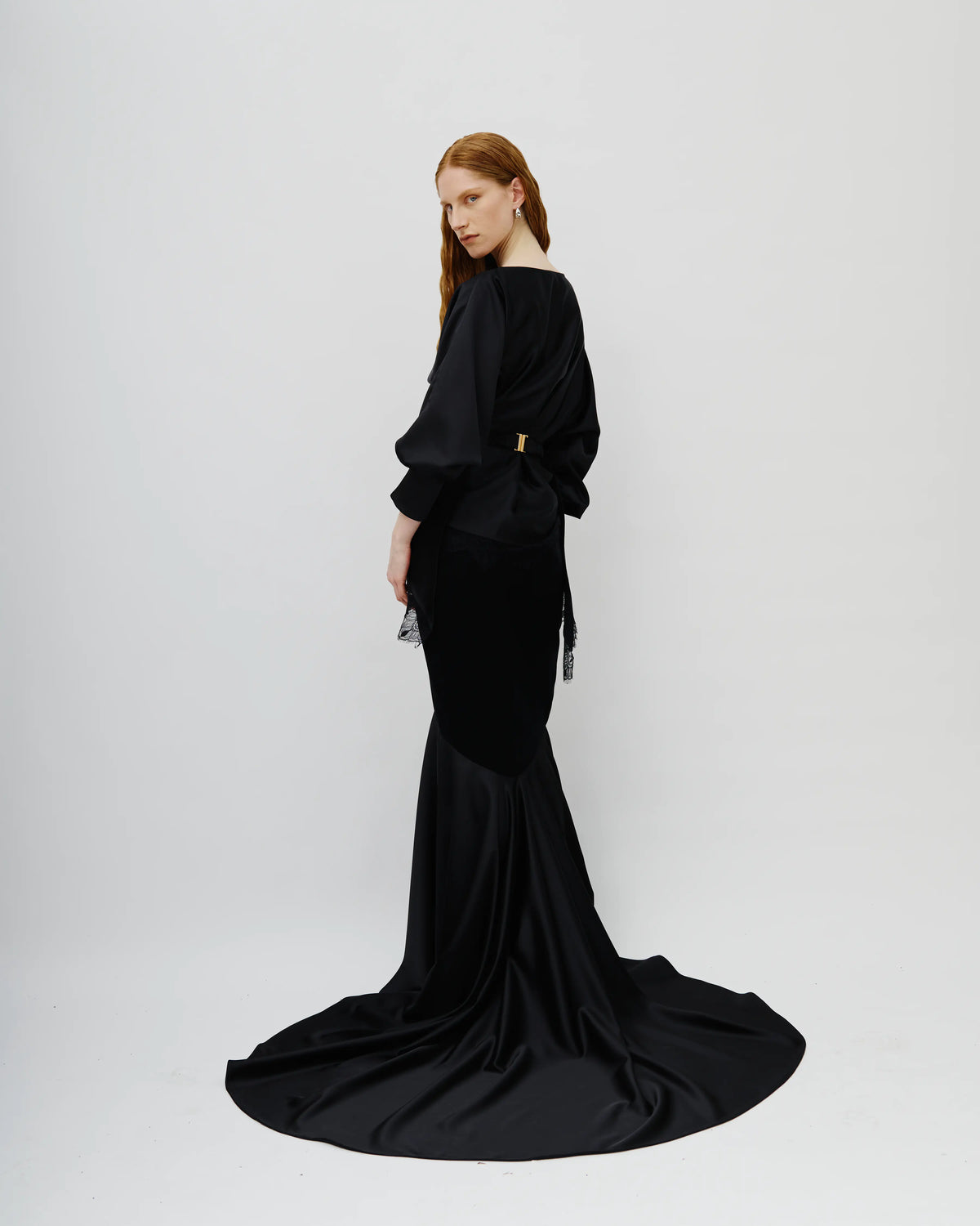 A model with red hair is shown from the back wearing a long black Avaro Figlio satin mermaid gown with a dramatic train against a light gray studio background.