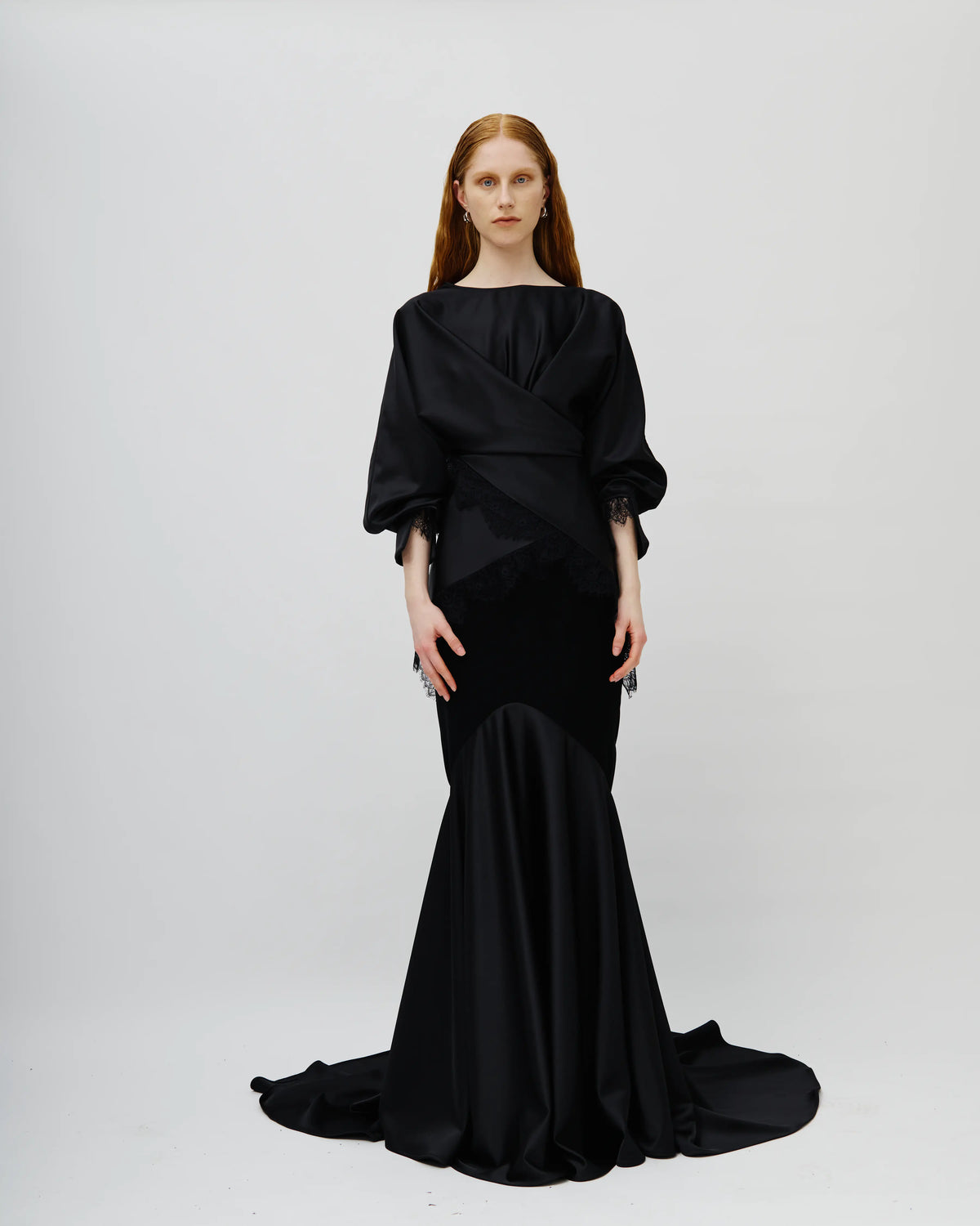 Model with red hair wearing a floor-length black Avaro Figlio gown with a draped bodice, voluminous elbow-length sleeves, and a satin mermaid-style skirt with a train, photographed against a light gray studio background.