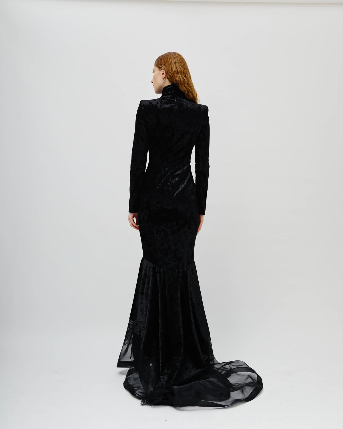 A woman with red hair is shown from the back wearing a long Avaro Figlio black velvet mermaid gown with a sheer train, photographed against a plain light gray studio background.