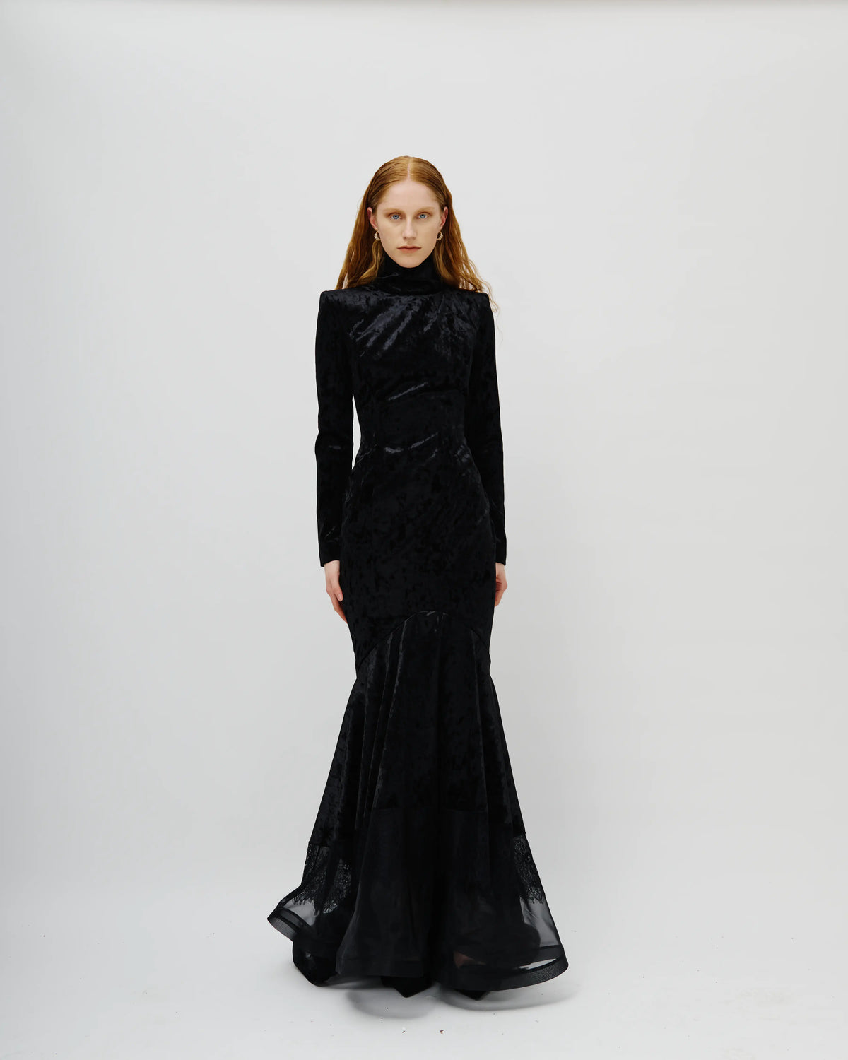 A model with red hair wearing an Avaro Figlio long-sleeved black velvet mermaid gown, standing against a plain light gray studio background.