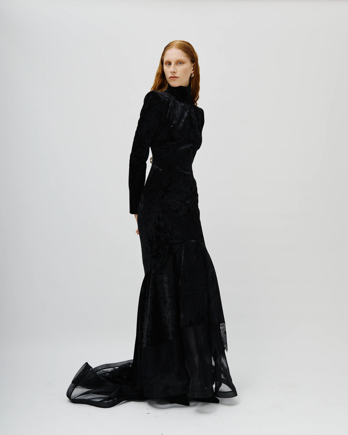 A model with red hair wearing a long-sleeved black Avaro Figlio velvet mermaid gown with a sheer tulle train, standing against a light gray studio background.